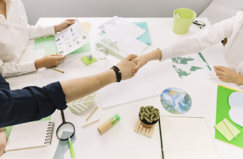 Two people shaking hands over a desk with business and environmental documents.