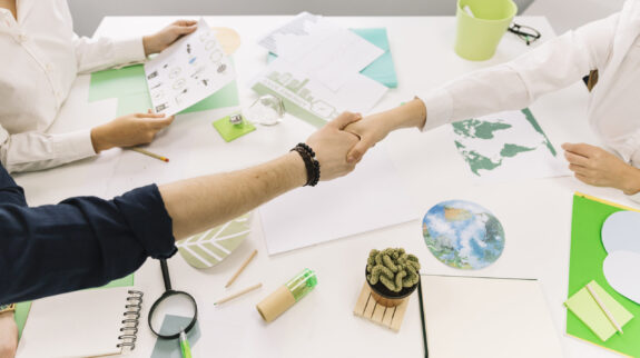 Two people shaking hands over a desk with business and environmental documents.